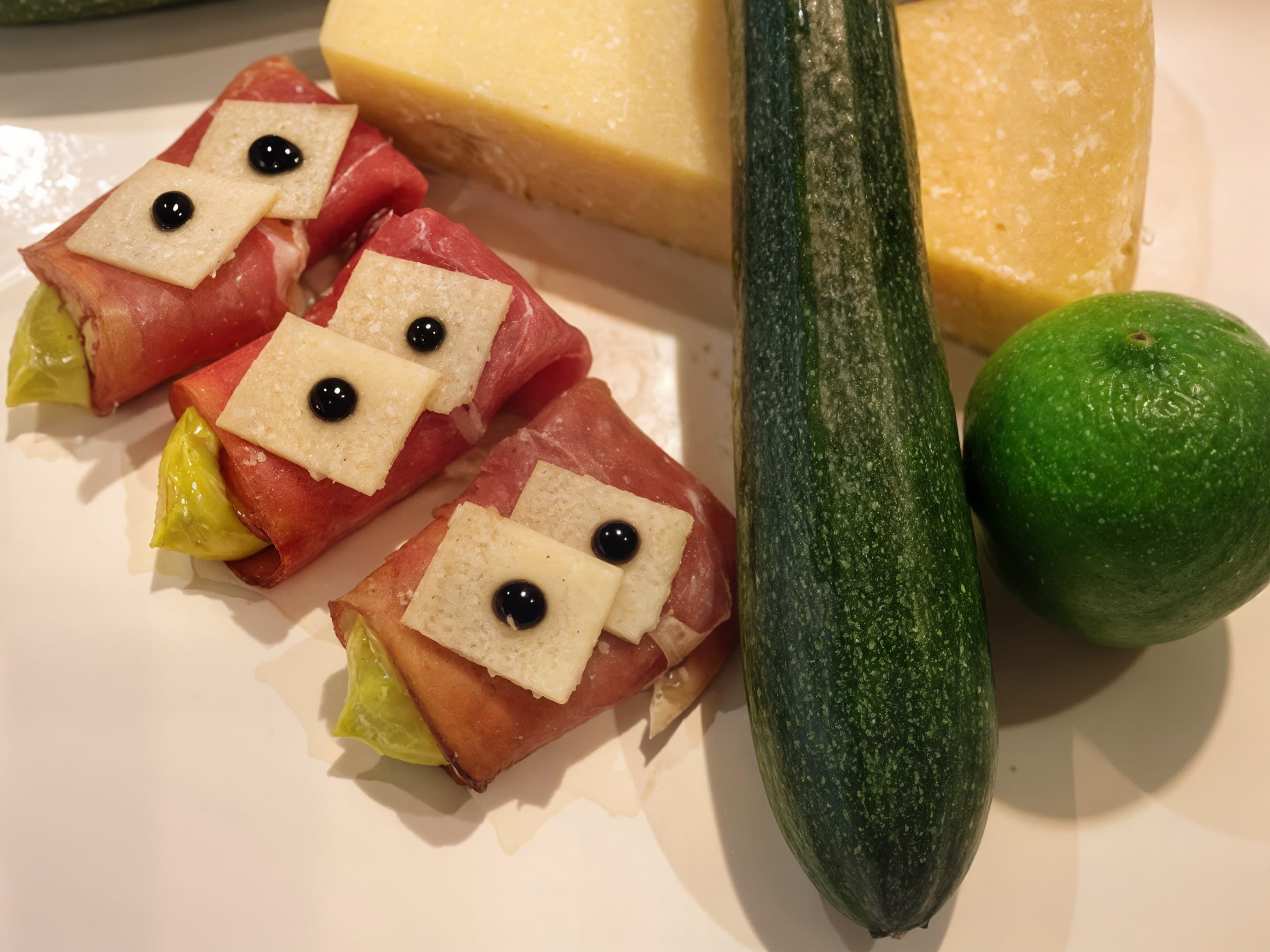 Schinkenröllchen mit Käsescheiben und Balsamico-Tupfen, daneben Zucchini, Käse und Limette