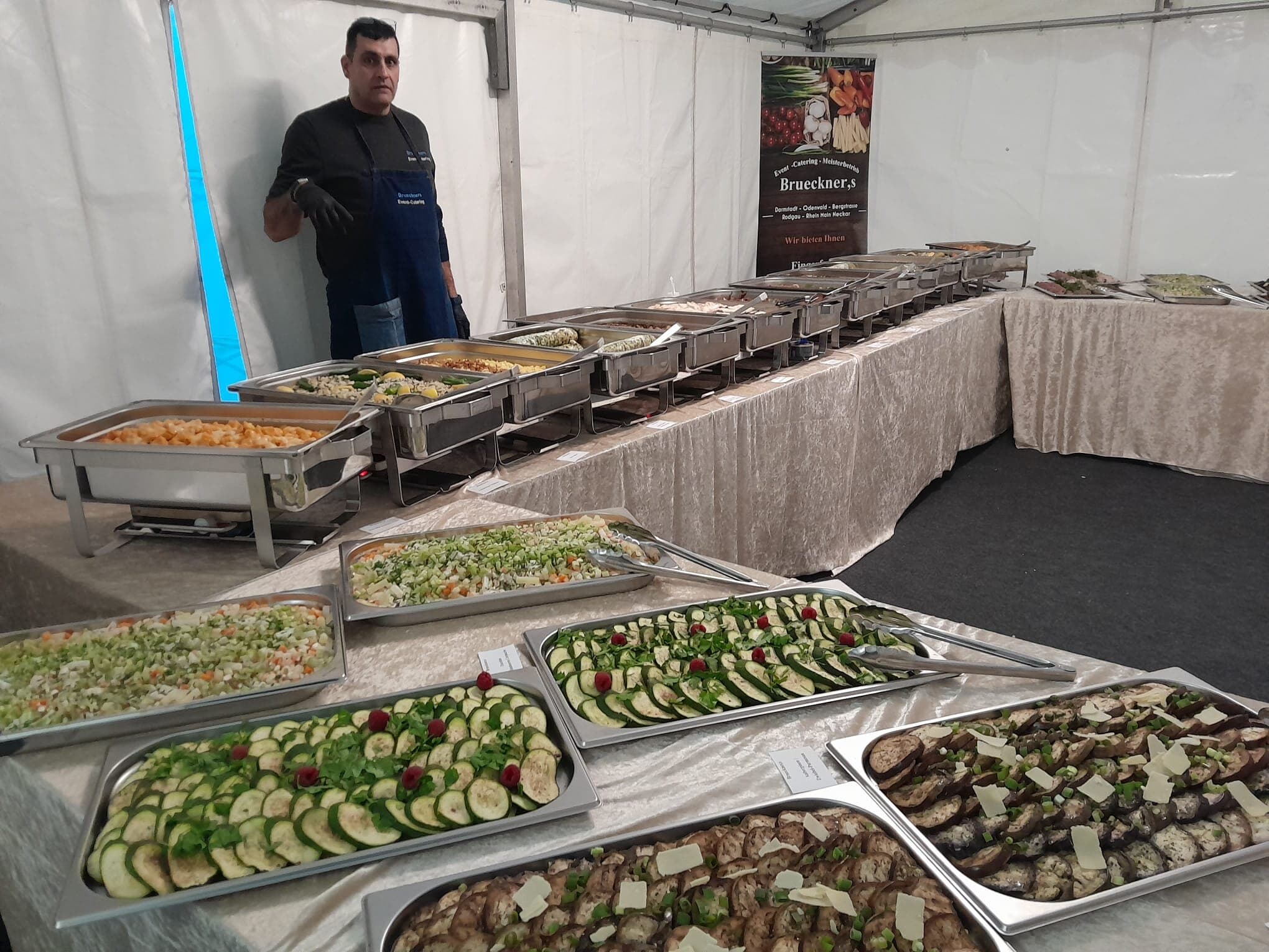 Warmes Buffet mit Warmhaltebehältern im Zelt, ein Mann in Schürze dahinter